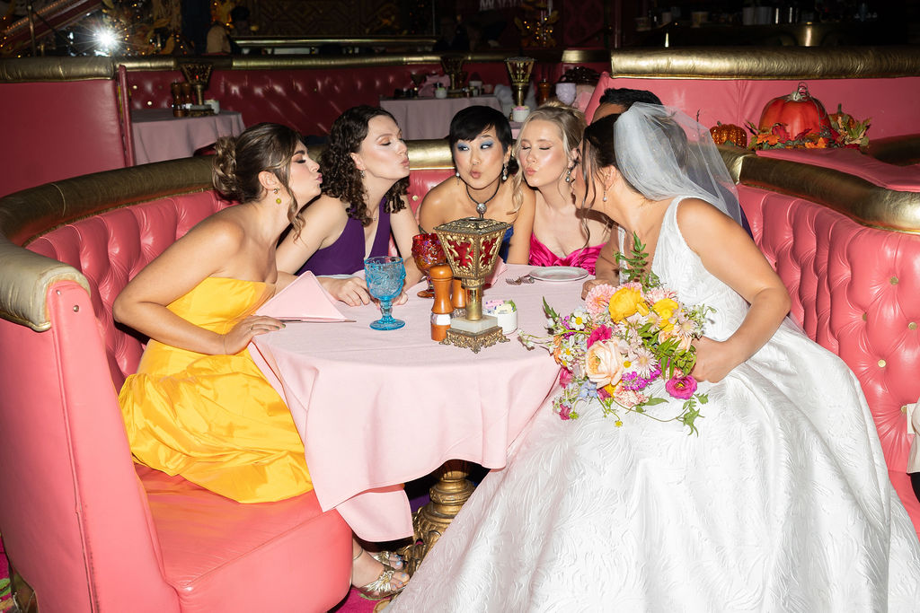 Bridesmaids and the bride make kissy faces around a pink booth table during her Madonna Inn wedding