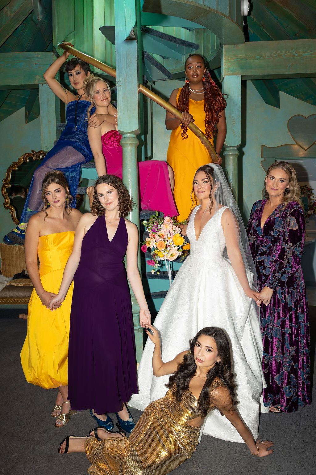 A bride and her bridesmaids in colorful gowns lounging around a spiral staircaseMadonna Inn wedding