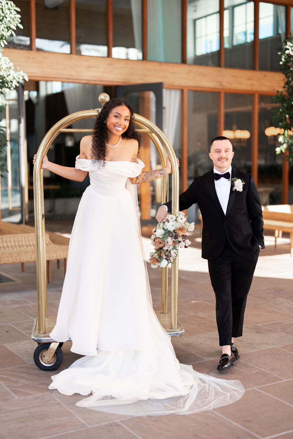 A bride and groom play on a luggage cart at the front entrance with big smiles