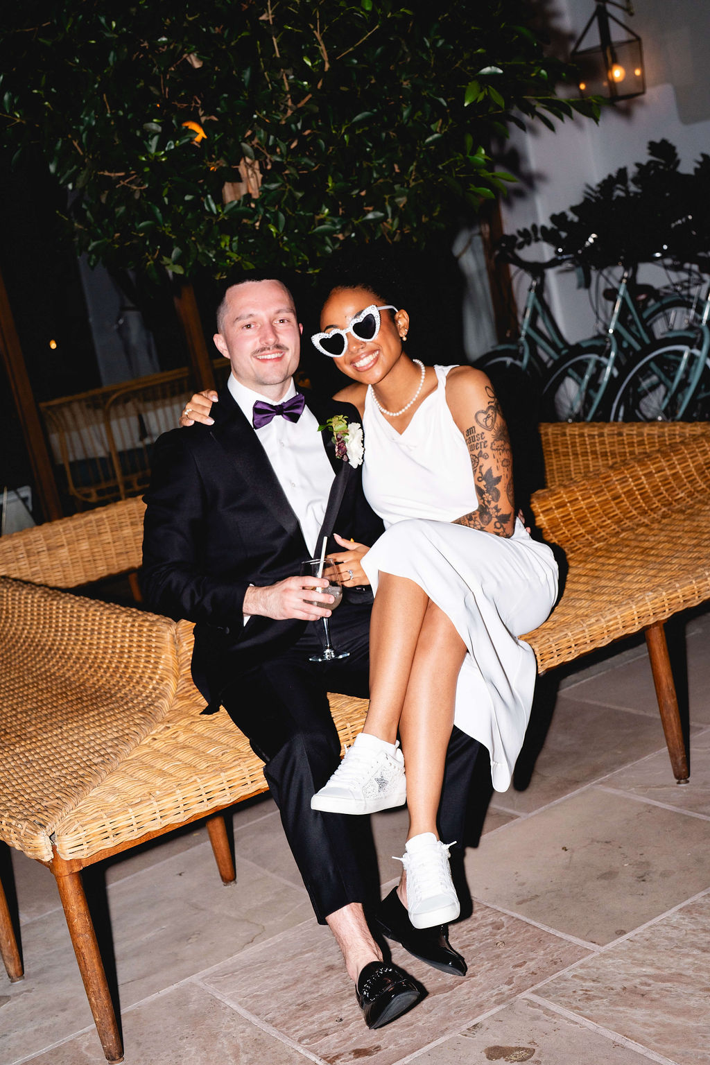 A bride smiles while sitting in the lap of her groom on a wicker bench