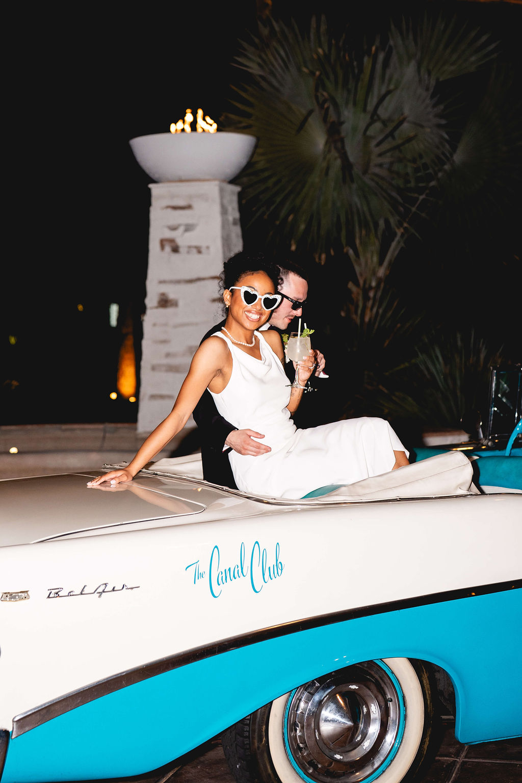 Happy newlyweds snuggle with drinks on the back bench of a vintage convertible